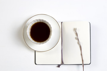 A white cup of coffee on a white notebook and a willow branch on a white wooden table.