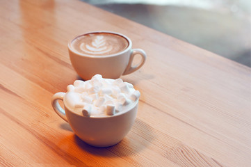 Two cups of cappuccino with latte art and marshmallow on wooden background. Beautiful foam, greenery ceramic cups, stylish toning, place for text.