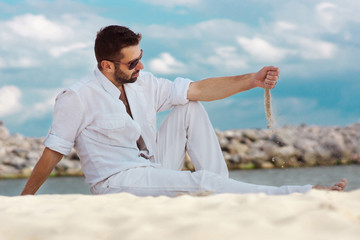 a man in white clothes and glasses sits on a white sand beach of a pond, ocean, sea, bay, lagoon