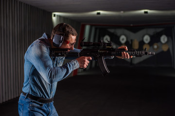 An adult man in jeans clothes, headphones and glasses, holding a Kalashnikov rifle with an optical...