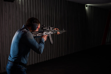 An adult man in jeans clothes, headphones and glasses, holding a Kalashnikov rifle with an optical sight. Trains fire with automatic weapons at the shooting range. - obrazy, fototapety, plakaty