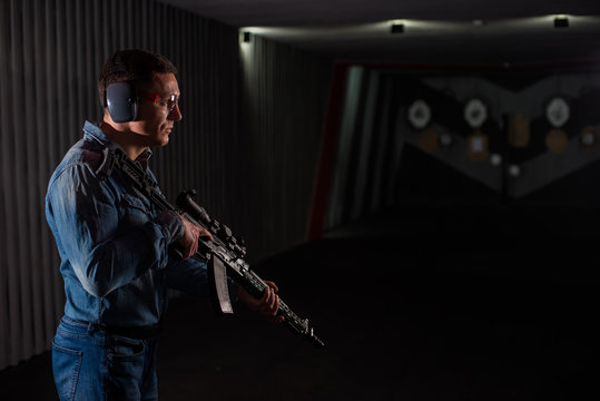 An adult man in jeans clothes, headphones and glasses, holding a Kalashnikov rifle with an optical sight. Trains fire with automatic weapons at the shooting range.