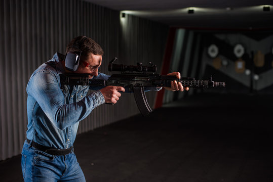 An adult man in jeans clothes, headphones and glasses, holding a Kalashnikov rifle with an optical sight. Trains fire with automatic weapons at the shooting range.