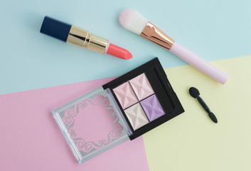 Flat lay of creative female decorative cosmetic for orange lipstick, pink cheek brush and eyeshadow palette on the colorful background with copy space