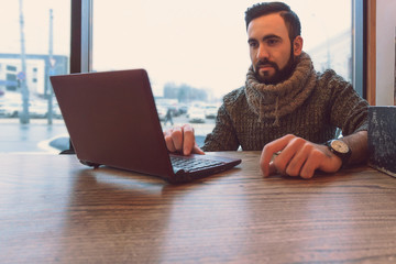 A young man with a beard working behind a laptop sitting in a cafe at a table with large panoramic windows