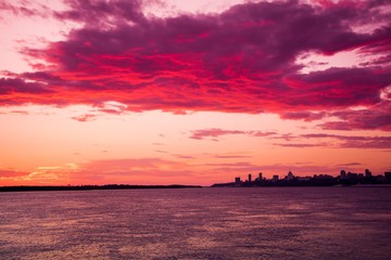 a city view on the horizon during the pink sunset with a river on a foreground and beautiful clouds