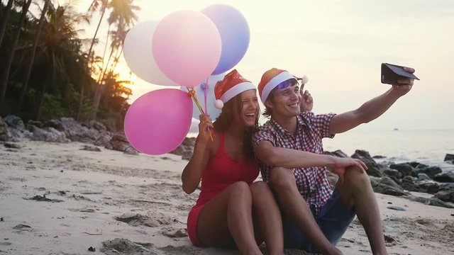 Happy funny couple sit on the beach with multicolored balloons on Christmas travel holidays taking selfie picture with smartphone wearing santa hat during amazing sunset. slow motion. 1920x1080