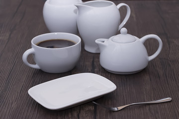 A white cup of coffee and other white utensils on brown wooden table. Breakfast.