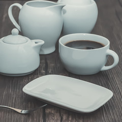 A white cup of coffee and other white utensils on brown wooden table. Breakfast.
