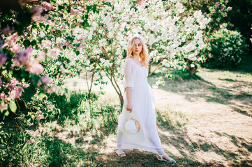 Fine art portrait of young blonde girl in white elegant dress in summer blooming lilac garden....