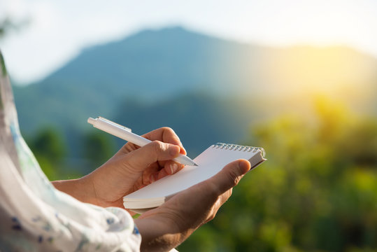 Hand writing on note pad.Woman hands holding  note pad writing with white pen  relaxing  outdoor at sunset ,blurred mountain background...