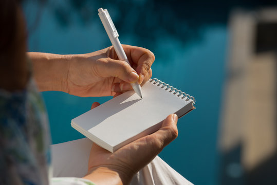 Hand writing on note pad.Woman hands holding  note pad writing with white pen relaxing by the pool at sunset ,blurred background.