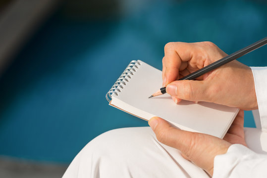 Hand writing on note pad.Woman hands holding  note pad writing with black pencil relaxing by the pool at sunset ,blurred background.