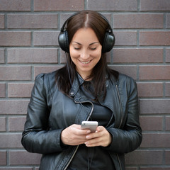 A beautiful girl in a black leather jacket listens to music from external headphones from her phone. Brick wall. On the street. Complicating with music. New album.