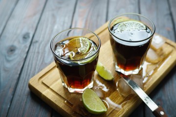 Cuba Libre cocktail with rum and cola served on wooden board with lime slices and melting ice cubes