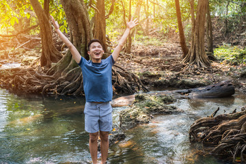 Relaxing for life natural therapy concept: Young man stands on rocks Stream river fast with raised arms up, smiling at forest for peachful. It show moisture tranquility, refreshing Travel of Thailand