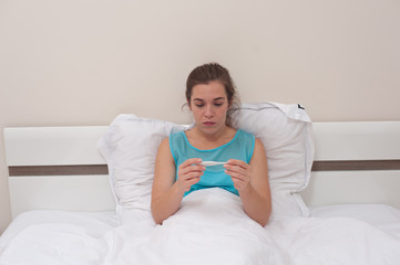 Disease and poor health. Portrait of Young woman in blue dress is sitting on the bed and holding thermometer in hands