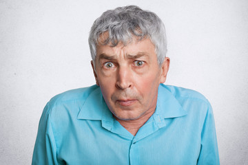 People, age and emotions concept. Surprised grey haired elderly man looks with unexpeted expression into camera, wears blue formal shirt, isolated over white background. Mature male poses indoor