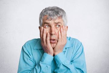 Photo of discontent frustrated grey haired male, keeps hands on cheeks, looks desperately somewhere, wears formal blue shirt, isolated over white concrete wall. People, problems and age concept
