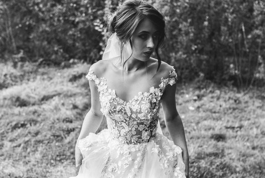 Beautiful bride with elegant simple hairdo is standing in the forest. Low open back white dress with flowers and tulle veil. Stylish hairstyle in outdoors romantic wedding photo. Black and white.