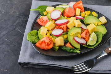 Fresh avocado salad with tomatoes, spinach, cheese, radish and corn in a black plate  on dark  background  Healthy food concept.