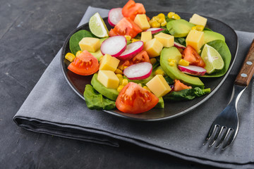 Fresh avocado salad with tomatoes, spinach, cheese, radish and corn in a black plate  on dark  background  Healthy food concept.