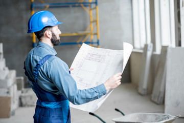 Builder or foreman in working uniform expertising the structure standing with blueprint at the construction site indoors