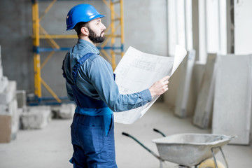 Builder or foreman in working uniform expertising the structure standing with blueprint at the construction site indoors