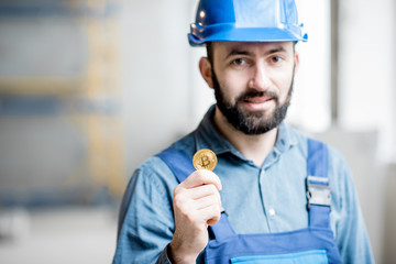 Builder in working uniform holding cryptocoin as a salary standing at the construction site indoors