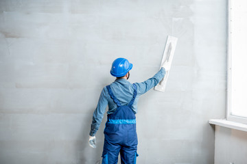 Plasterer in blue working uniform plastering the wall indoors - obrazy, fototapety, plakaty