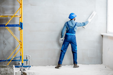 Plasterer in blue working uniform plastering the wall indoors - obrazy, fototapety, plakaty