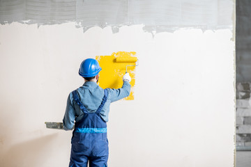 Workman in uniform painting wall with yellow paint at the construction site indoors