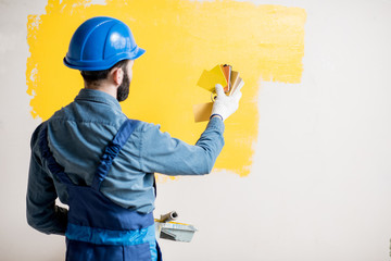 Painter in blue workwear comparing yellow painted wall with color swatches indoors