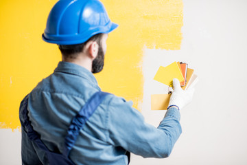 Painter in blue workwear comparing yellow painted wall with color swatches indoors