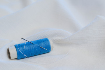 Fototapeta na wymiar Blue thread spooll with sewing needle on white cotton cloth