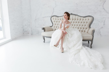 Bride looking out the window, she waits for the groom sitting on big classic sofa. Minimalistic loft studio interior