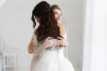 Fototapeta na wymiar luxury bride hugging bridesmaid and smiling, joyful moment in minimalistic loft white brick background