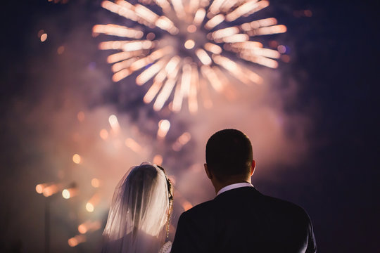 Happy hugging bride and groom watching beautiful colorful fireworks night sky