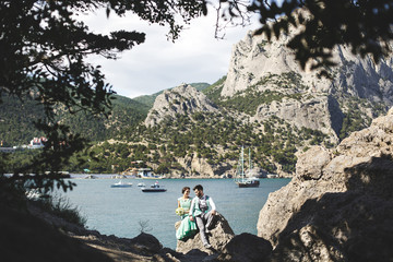The bride and groom on nature in the mountains near the water. Suit and dress color Tiffany. Sitting on the stone.
