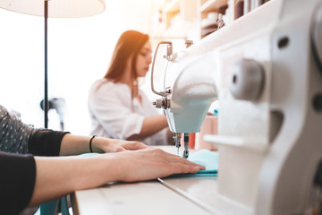 Creative dressmaker woman working with cloth on sewing machine. Two seamstress in design bureau making new clothing concept. Small business. Flare effect. Blurred background