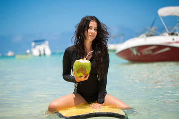 Portrait of a beautiful surfing girl with green coconut from palm tree sit on yellow surf longboard...