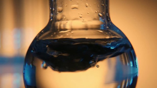 A macro shot of transparent water in a round bottom flask shaken in a slightly lit medical laboratory. The background is dark orange.