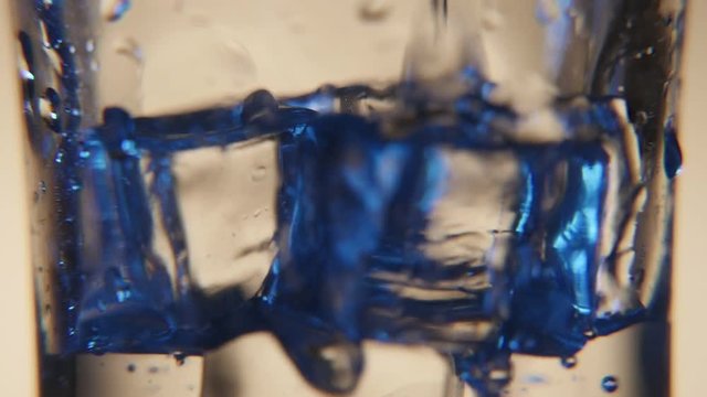 An impressive macro shot of a transparent glass full of bluish ice cubes in a bar. Water stream with shining bubble is poured in the glass 