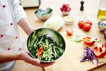 Metallic bowl with cut vegetables held by chef on background of workplace
