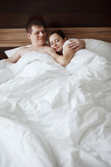 Fototapeta premium Young beautiful husband and wife resting in bed under a white blanket in a hotel room