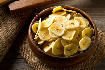 Heap of dried banana chips snack in wooden bowl on dark table