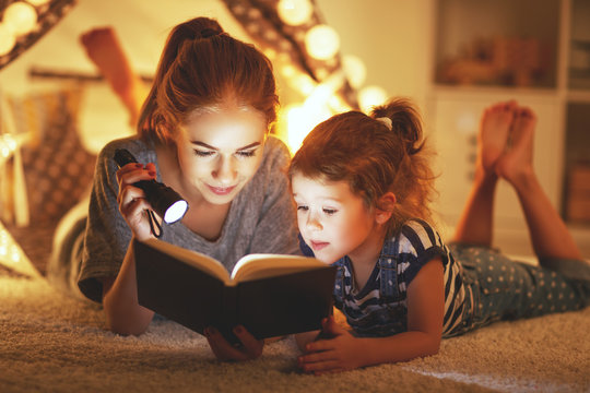 mother and child daughter reading a book and a flashlight before going to bed