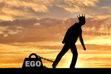 A big weight in the form of an ego is chained to the foot of a selfish and narcissistic man with a crown on his head