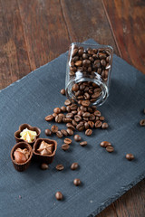 Side view of overturned glass jar with coffee beans and chocolate candies on wooden background, selective focus