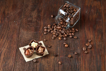 Side view of overturned glass jar with coffee beans and chocolate candies on wooden background, selective focus